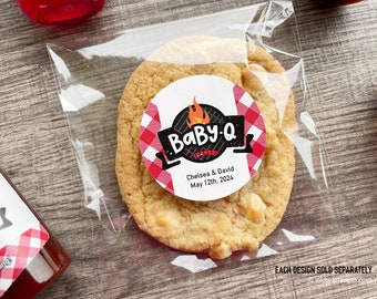 May include: A white cookie with white chocolate chips in a clear plastic bag with a round sticker that says "BaBy-Q" with a red and black checkered pattern and the text "Chelsea & David May 12th, 2024".