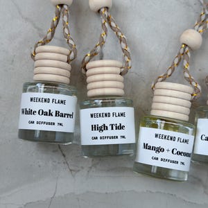 May include: Three small glass car diffuser bottles with light wood caps and braided cords featuring gold metallic thread. Each bottle has a white label reading WEEKEND FLAME, the scent name, and CAR DIFFUSER 7ML. Scents include White Oak Barrel, High Tide, and Mango + Coconut.