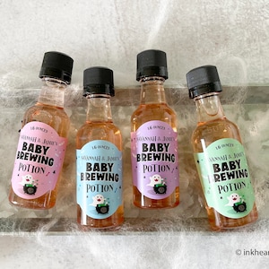 May include: Four small bottles of liquid with black caps, each labeled "Baby Brewing Potion" with a ghost and cauldron graphic. The labels are in pink, blue, and green. The bottles are on a surface with spiderweb decor.