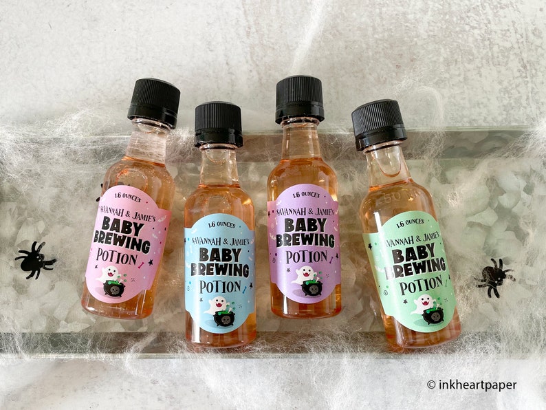 May include: Four small bottles of liquid with black caps, each labeled "Baby Brewing Potion" with a ghost and cauldron graphic. The labels are in pink, blue, and green. The bottles are on a surface with spiderweb decor.
