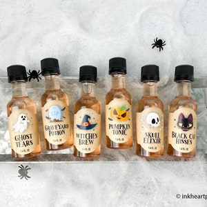 May include: Six small glass bottles with black lids and Halloween-themed labels. The labels feature illustrations of a ghost, a graveyard, a witch's hat, a pumpkin, a skull, and a black cat. The bottles are arranged on a white surface with a spiderweb pattern.