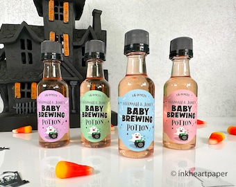Op de afbeelding: Vier kleine glazen flesjes met etiketten waarop "Savannah & Jamie's Baby Brewing Potion" staat in zwarte tekst op een roze, blauwe, groene en lichtroze achtergrond. Elk flesje heeft een zwarte dop en een spookillustratie. De flesjes zijn in een rij op een wit oppervlak geplaatst met verspreide maïs snoepjes.