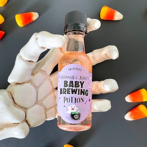 May include: A small glass bottle with a black cap, labeled "Savannah & Jamie's Baby Brewing Potion." The label features a ghost and a cauldron. Candy corn is scattered around the bottle, which is held by a white skeleton hand. The bottle contains 1.6 ounces of liquid.