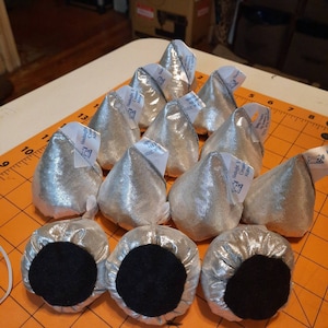 May include: A group of 12 silver fabric fortune cookie-shaped pouches with black felt bottoms. Each pouch has a white label with blue text that reads "Hindsight Creative Abby" and "Est. 2020".