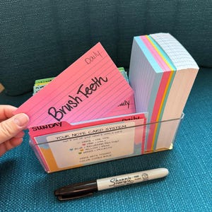 May include: A clear plastic organizer box containing a stack of multi-colored index cards and a set of tabbed dividers. One pink card is held up, displaying the handwritten text "Brush Teeth" and "Daily". A black Sharpie marker lies in front.
