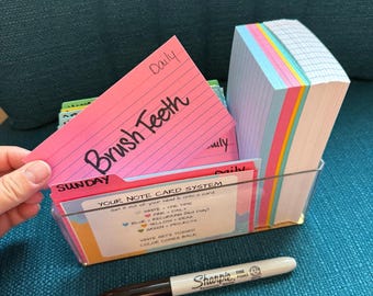 May include: A clear plastic organizer box containing a stack of multi-colored index cards and a set of tabbed dividers. One pink card is held up, displaying the handwritten text "Brush Teeth" and "Daily". A black Sharpie marker lies in front.