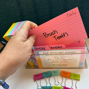 May include: A clear plastic organizer box containing colorful index cards, including a pink card labeled "Brush Teeth" and "Daily". A set of metal binder clips with labels for days of the week and tasks like "Today", "Tomorrow", "TO DO", and "Urgent" are arranged below.
