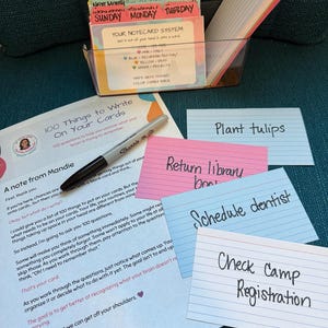 May include: A notecard system kit featuring a clear plastic organizer filled with color-coded index cards, a black marker, and a printed guide titled 100 Things to Write On Your Cards. Several lined index cards with handwritten tasks are displayed.