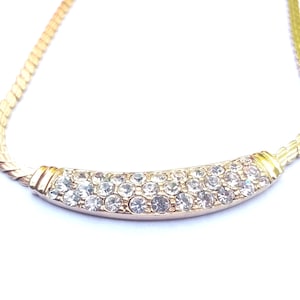 May include: A gold-toned necklace with a curved bar pendant. The pendant is encrusted with numerous clear, sparkling crystals. The chain has a woven design. The necklace is set against a white background.