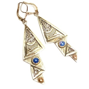 Pode incluir: Um par de brincos prateados com um design Art Déco. Cada brinco apresenta uma forma triangular com intrincados padrões de filigrana e geométricos, acentuados com pedras azuis e cor de cobre. Os brincos têm fechos de alavanca.