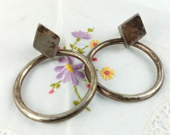 May include: A pair of silver-toned hoop earrings with a square accent. The earrings are circular with a square shape at the top. The earrings are set against a white background with embroidered flowers.