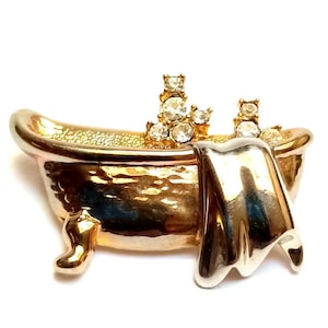May include: A gold-toned bathtub-shaped brooch with a silver-colored towel draped over the side. The bathtub has three small legs and is adorned with clear, sparkling crystals. A unique accessory.