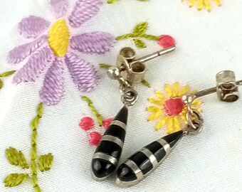 May include: A pair of silver dangle earrings with black and silver striped teardrop-shaped pendants. The earrings are displayed on a white embroidered surface with purple and yellow floral designs.