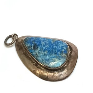 Puede incluir: Colgante plateado con una piedra azul. La piedra, de forma irregular, presenta una mezcla de tonos azules. El colgante tiene un pequeño lazo en la parte superior para una cadena.