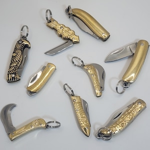 May include: A collection of ten small, decorative pocket knives with gold-toned metal handles and blades. The knives have various designs, including a heart, a bird, and a fish.
