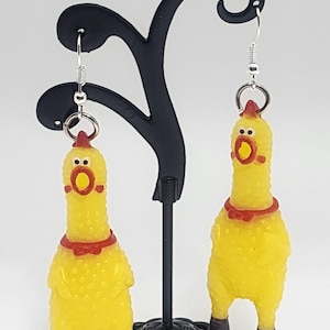 May include: A pair of yellow rubber chicken earrings with red bows. The earrings are hanging from a black earring stand.