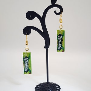 May include: A pair of gold dangle earrings with green and white Doublemint gum wrappers. The earrings are hanging on a black earring stand.