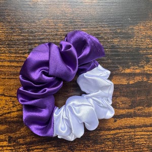 May include: A two-tone scrunchie, half purple and half white, made of a shiny satin-like fabric. The hair accessory is circular and sits on a wooden surface. The scrunchie is a hair accessory.