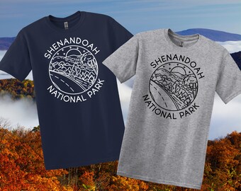 May include: Two short-sleeved t-shirts, one navy blue and one grey, each with a circular graphic. The graphic reads "Shenandoah National Park" around a road, trees, and sun design. The shirts are set against a backdrop of autumn foliage.