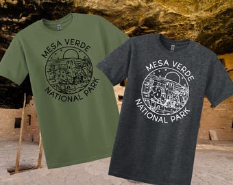 May include: Two t-shirts, one olive green and one charcoal grey, each with a circular graphic. The graphic reads "Mesa Verde National Park" and shows a landscape with buildings and a sun.