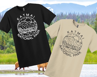 May include: Two t-shirts, one black and one beige, each with a circular graphic that reads "Katmai National Park." The graphic features a mountain range, sun, and stars. The shirts are short-sleeved.