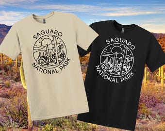 May include: Two t-shirts, one beige and one black, with a circular graphic that reads "Saguaro National Park." The design includes a sun, cacti, and mountains. The shirts have short sleeves.