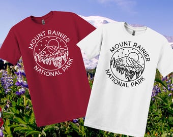 May include: Two t-shirts, one red and one white, each with a circular graphic reading "Mount Rainier National Park." The design includes a mountain, sun, and trees. The shirts are shown against a backdrop of a mountain and flowers.