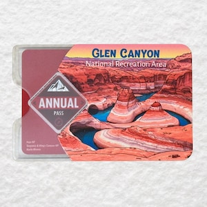 May include: A clear plastic holder containing a red and pink Glen Canyon National Recreation Area annual pass. The pass features a landscape image of the canyon with blue water and red rock formations. The pass also has the words "Annual Pass".