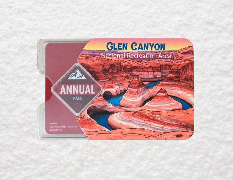 May include: A clear plastic holder containing a red and pink Glen Canyon National Recreation Area annual pass. The pass features a landscape image of the canyon with blue water and red rock formations. The pass also has the words "Annual Pass".