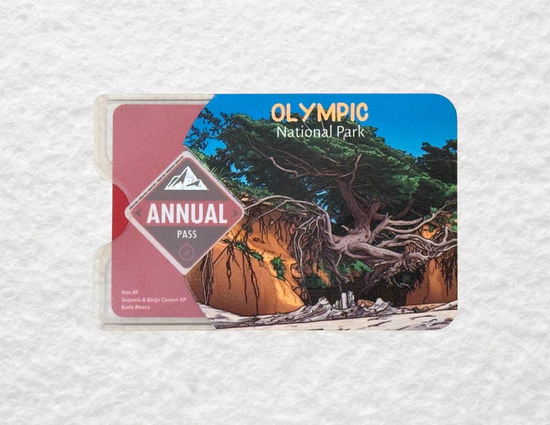 May include: An annual pass for Olympic National Park. The pass is in a clear plastic holder. The pass has a blue and red design with the words "Olympic National Park" and "Annual Pass". The pass features a landscape image of a tree.