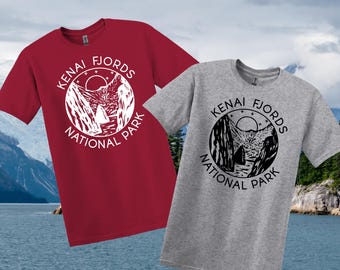 May include: Two short-sleeved t-shirts, one red and one grey, each with a circular graphic. The graphic shows the text "KENAI FJORDS NATIONAL PARK" around a mountain and water scene.