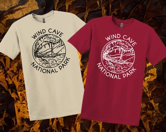 May include: Two short-sleeved t-shirts, one cream and one burgundy, are shown. Each has a circular graphic with the text "Wind Cave National Park" and a detailed illustration of a cave and landscape.