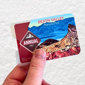 May include: A Death Valley National Park annual pass card is held in a clear plastic holder. The card features a scenic landscape of the park with blue skies and mountains. The words "DEATH VALLEY National Park" are printed at the top.