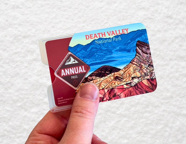 May include: A Death Valley National Park annual pass card is held in a clear plastic holder. The card features a scenic landscape of the park with blue skies and mountains. The words "DEATH VALLEY National Park" are printed at the top.