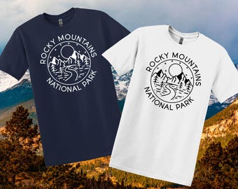 May include: Two t-shirts, one navy blue and one white, each with a circular graphic. The graphic displays the text "Rocky Mountains National Park" around a design of mountains, a river, trees, and a moon. The shirts are short-sleeved.
