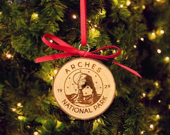May include: A wooden Christmas bauble with a red ribbon. The bauble is a round slice of wood with the words "Arches National Park" and a depiction of Delicate Arch. The bauble is hanging on a Christmas tree.