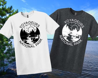 May include: Two t-shirts, one white and one charcoal grey, each with a circular graphic reading "VOYAGEURS NATIONAL PARK." The graphic shows a landscape with trees, water, and a moon. The shirts are short-sleeved.