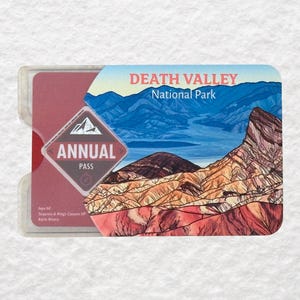 May include: A Death Valley National Park annual pass in a clear plastic holder. The pass features a landscape illustration of mountains and desert scenery. Text on the pass includes "ANNUAL PASS" and "DEATH VALLEY National Park."
