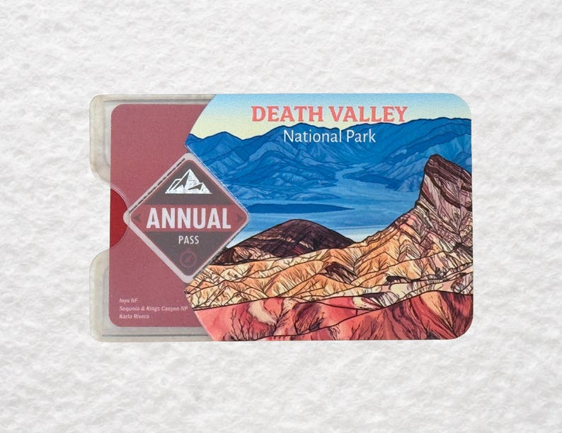 May include: A Death Valley National Park annual pass in a clear plastic holder. The pass features a landscape illustration of mountains and desert scenery. Text on the pass includes "ANNUAL PASS" and "DEATH VALLEY National Park."