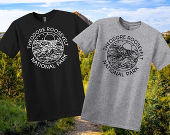 May include: Two t-shirts, one black and one grey, displaying a circular graphic with the text "Theodore Roosevelt National Park." The design includes a mountain range, sun, and stars. The shirts are short-sleeved.
