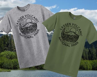 May include: Two t-shirts, one light grey and one olive green, each with a black circular graphic. The graphic reads "Lassen Volcanic National Park" and depicts a mountain scene with a sun and stars.