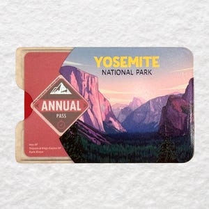 May include: Yosemite National Park annual pass with a scenic landscape illustration. The pass features a red and cream-colored holder, with the words "Yosemite National Park" in yellow. The pass itself has a mountain range illustration.