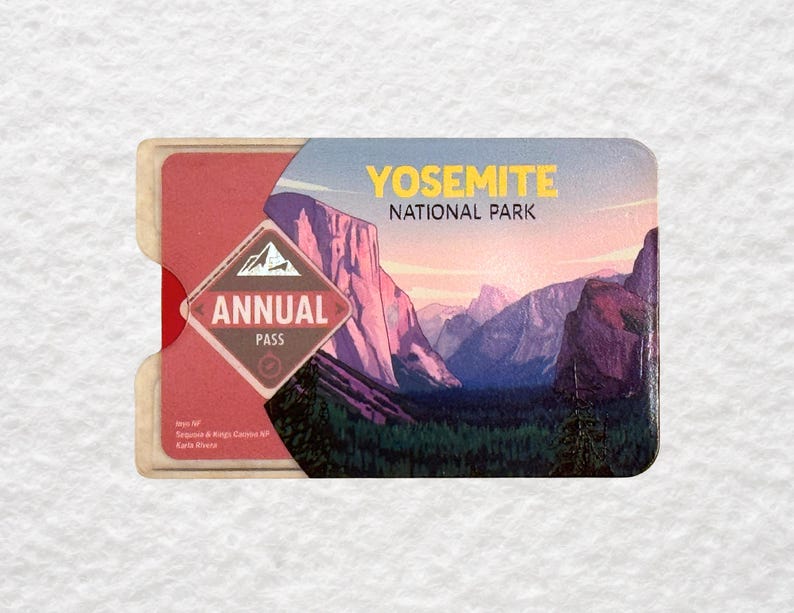 May include: Yosemite National Park annual pass with a scenic landscape illustration. The pass features a red and cream-colored holder, with the words "Yosemite National Park" in yellow. The pass itself has a mountain range illustration.