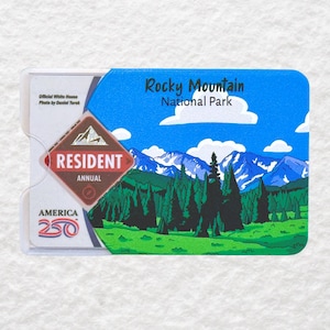May include: A Rocky Mountain National Park resident annual pass card. The card features a scenic illustration of mountains, trees, and a blue sky. The card is in a clear plastic holder with text that reads "America 250".