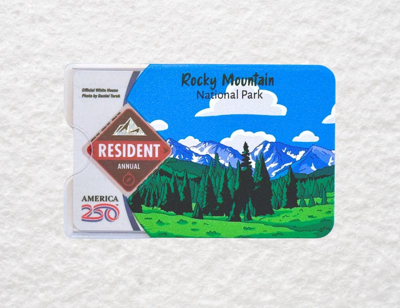 May include: A Rocky Mountain National Park resident annual pass card. The card features a scenic illustration of mountains, trees, and a blue sky. The card is in a clear plastic holder with text that reads "America 250".
