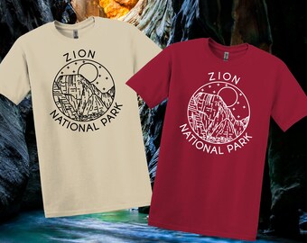 May include: Two short-sleeved t-shirts, one cream and one claret, each with a circular graphic reading "ZION NATIONAL PARK." The graphic shows a mountain scene, a sun, and stars. The shirts are set against a blurred background.