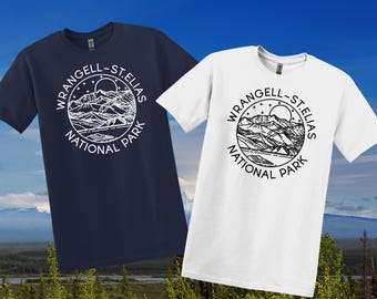 May include: Two short-sleeved t-shirts, one navy blue and one white. Each shirt displays a circular graphic with the text "WRANGELL-ST. ELIAS NATIONAL PARK" around a mountain and sun illustration. The shirts are made of a soft material.