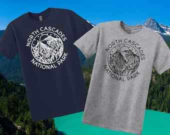May include: Two short-sleeved t-shirts, one navy blue and one grey, each with a circular graphic. The graphic reads "North Cascades National Park" around a mountain and sun design. The shirts are displayed against a backdrop of mountains and water.