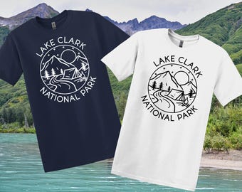 May include: Two t-shirts, one navy blue and one white, each displaying a circular graphic with the words "Lake Clark National Park." The design includes mountains, a sun, trees, a road, and a tent. The shirts are short-sleeved.