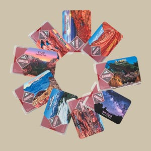 May include: A collection of annual National Park passes, each featuring a different park scene. The passes are in clear plastic holders and display park names like Zion, Arches, and Olympic. The word "ANNUAL" is printed on each pass.